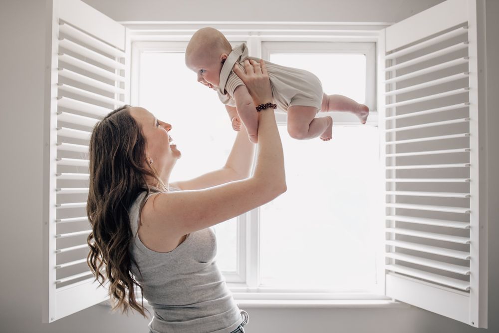 A mother throws her 4-month-old baby up in the air in front of a shuttered window.
