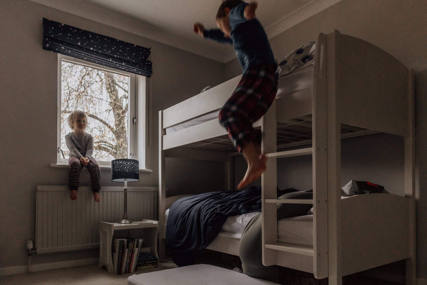 One boy jumps off the top bunk of some bunkbeds while the other watches from a window sill.