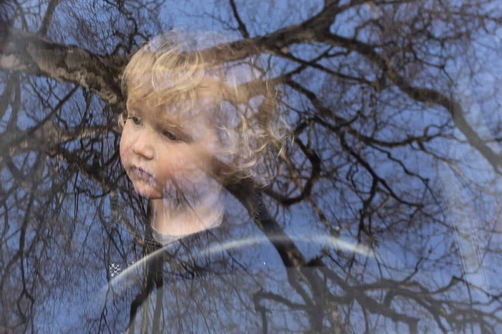 A young boy plays at driving in his parents' car, the reflection of a tree in the windscreen creates a double exposure effect.