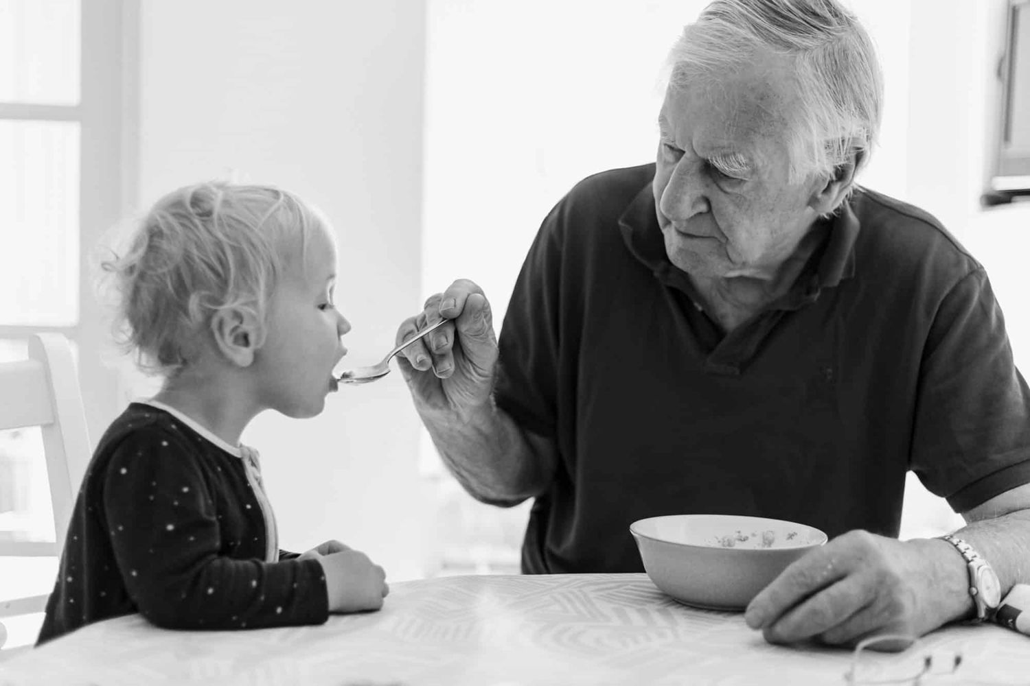 A grandfather feeds his grandchild, holding a spoon out towards his mouth.