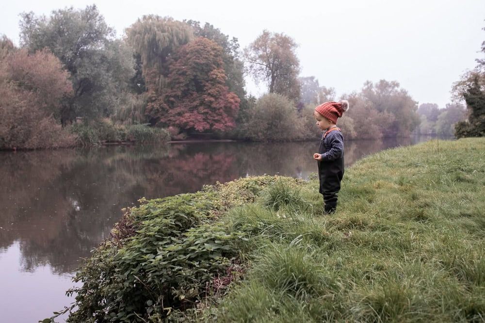 A child stands on the bank of the thames at Dorney wearing a bobble hat.