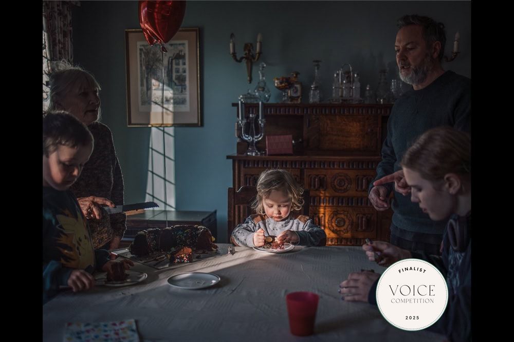 A boy sits at the head of a dining table tucking under his birthday cake.  He is surrounded by close family.