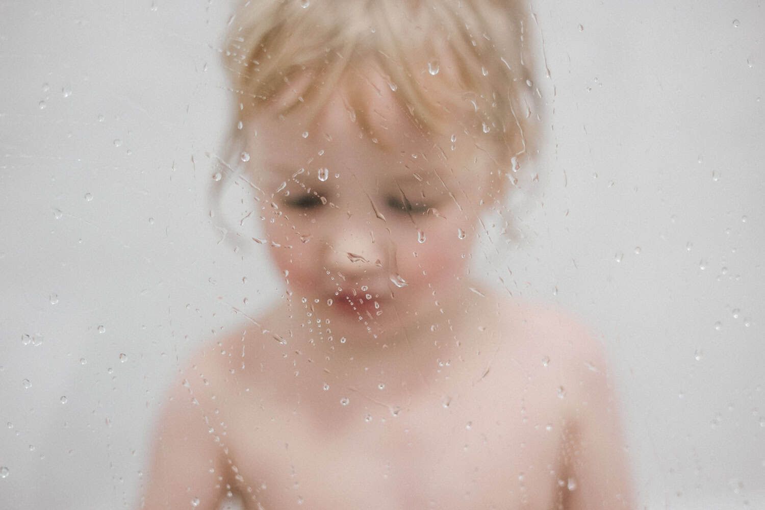 A young child in the bath, the image is taken through a shower screen so splashes of water form bokeh in front of his face.