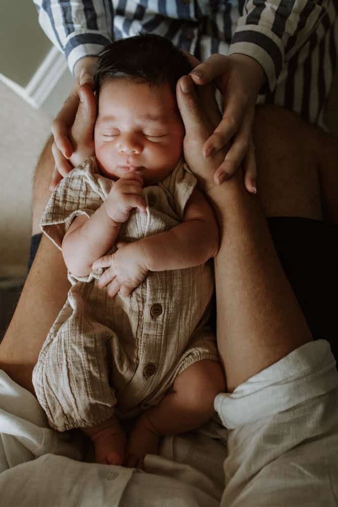 A mother's hands reach over a father's as they both craddle their newborn son.