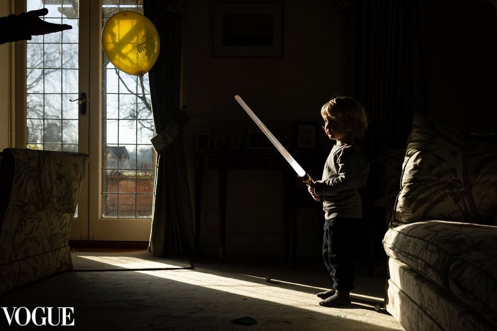 A boy plays indoor cricket with a lightsaber toy and a balloon.  The photo carries a stamp from Photo Vogue.