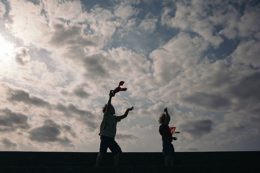 Two boys playing with their beach toys, one flings his arms out against the wind.