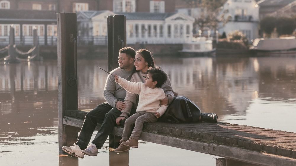 Parents sit with their toddler on a jetty over the river opposite the Compleat Angler in Marlow.