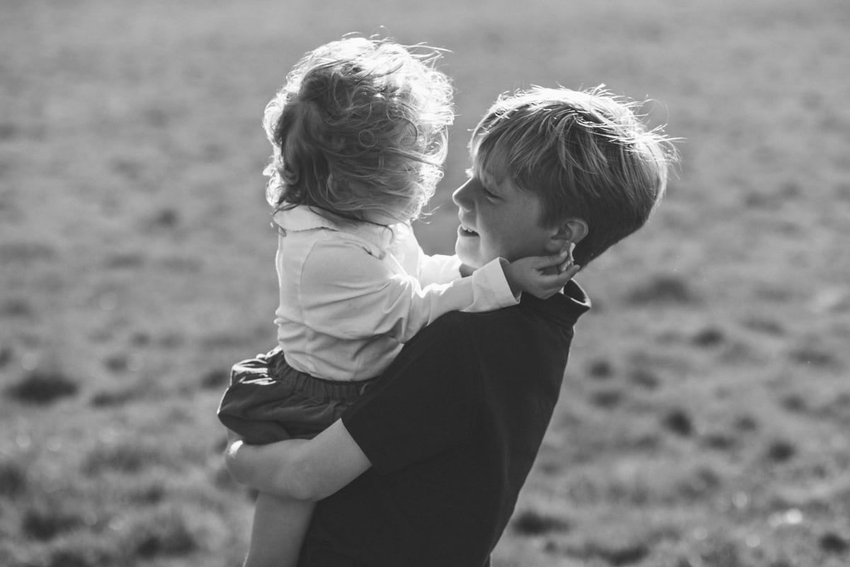 A boy holds his little sister in his arms, her hair blows over her face.
