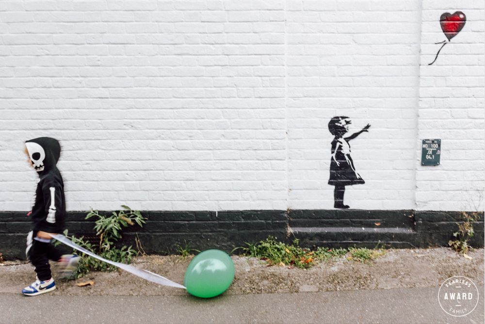 A young boy dressed as a skeleton and pulling a green balloon walks past a Banksy-inspired graffiti of a girl with a red balloon.