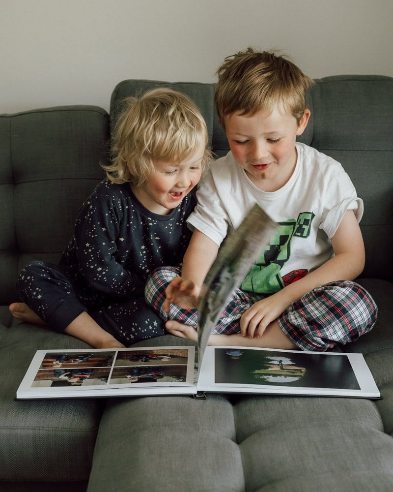 Two boys flick through a photobook together.  They laugh as they look through the memories it contains.