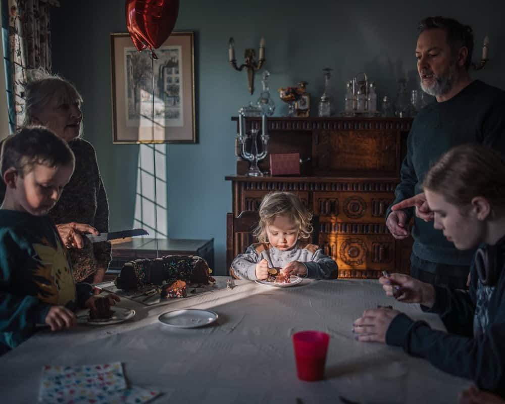 A small family gathering for a birthday party, the birthday boy eats a big slide of cake at the centre of the image.