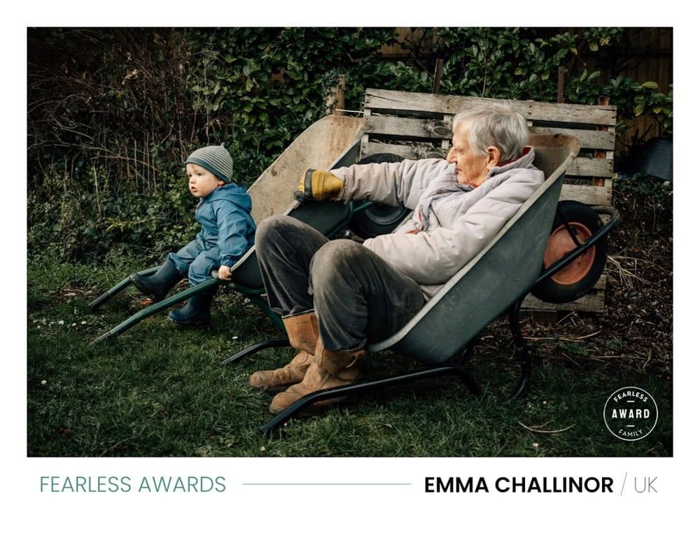 A grandfather sitting in a wheelbarrow holds another still for his grandchild to climb in.