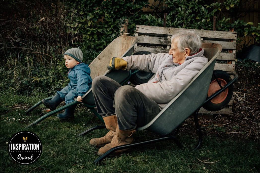 A grandfather holds a wheelbarrow still for his grandson to sit inside, next to him, on a break from gardening.