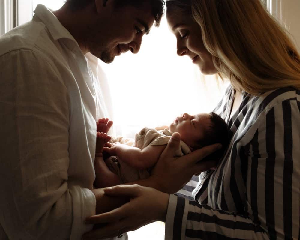 A mother and father cradle their newborn in this back-lit portrait.