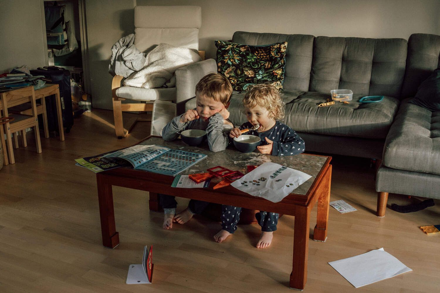 Two boys eating breakfast sharing a seat at a coffee table in a messy room.