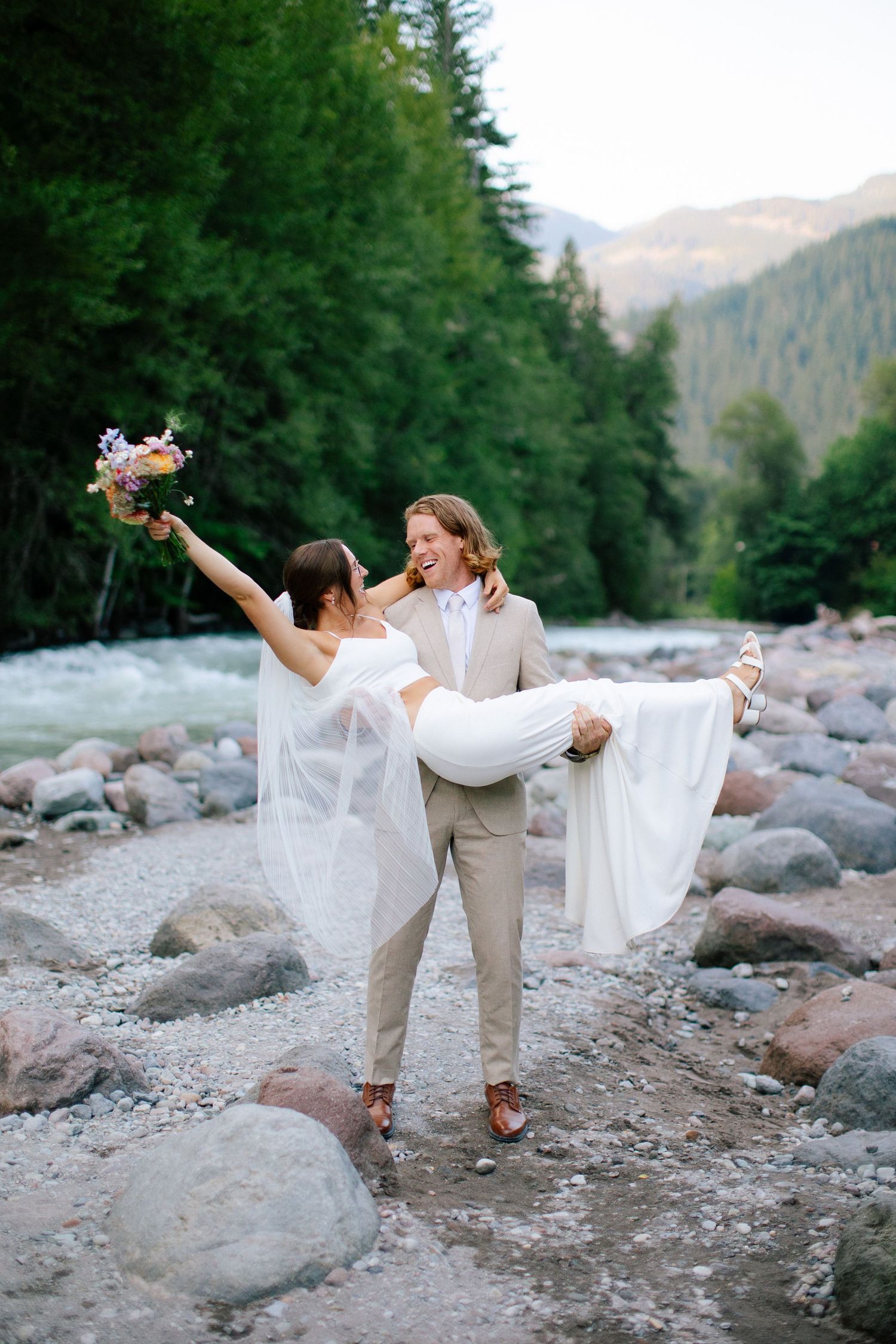 Bride and groom standing on rocky riverbank in Squamish, surrounded by forest and rushing water.