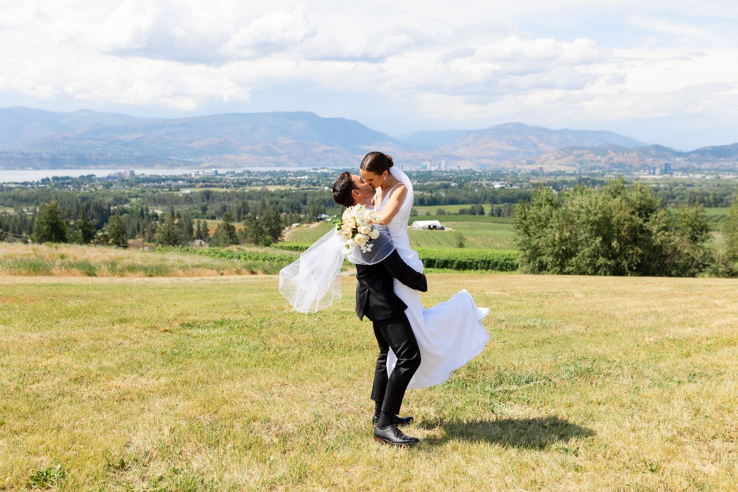 Groom lifting bride in open field with mountains behind.