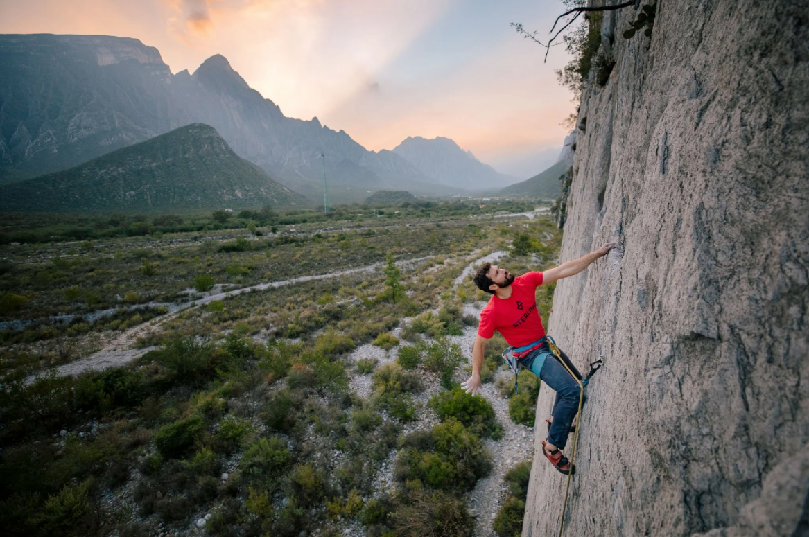Rock climber scaling vertical wall above valley at golden hour.