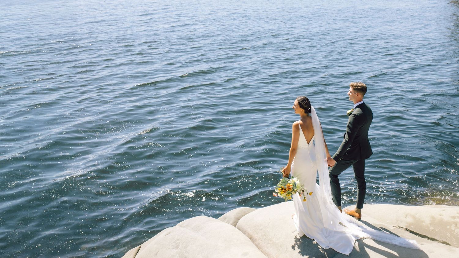 Bride and groom standing on rocky ocean shoreline.