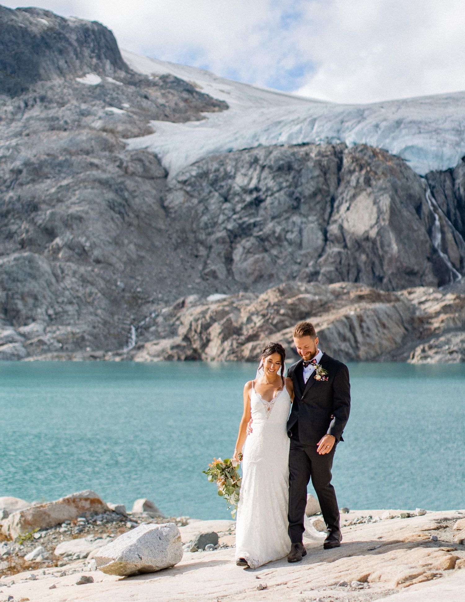 Couple embracing on rocky shoreline with dramatic coastal mountains behind them.