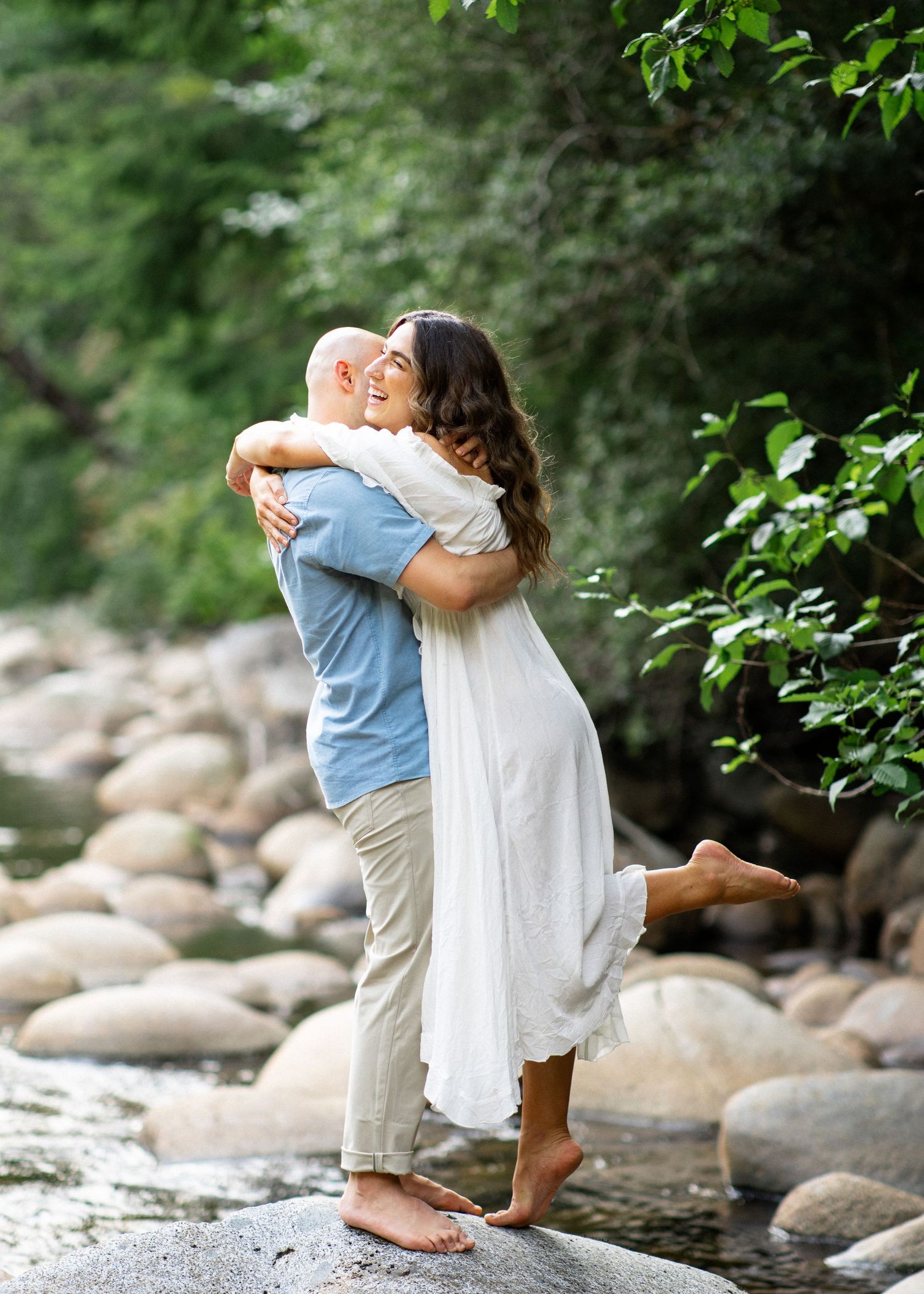 Bride and groom embracing on river rocks in forest setting.