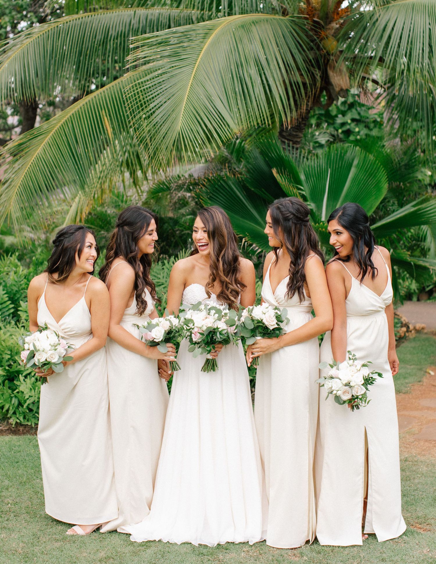 Bridesmaids in sage green dresses standing beneath palm leaves at a summer wedding.