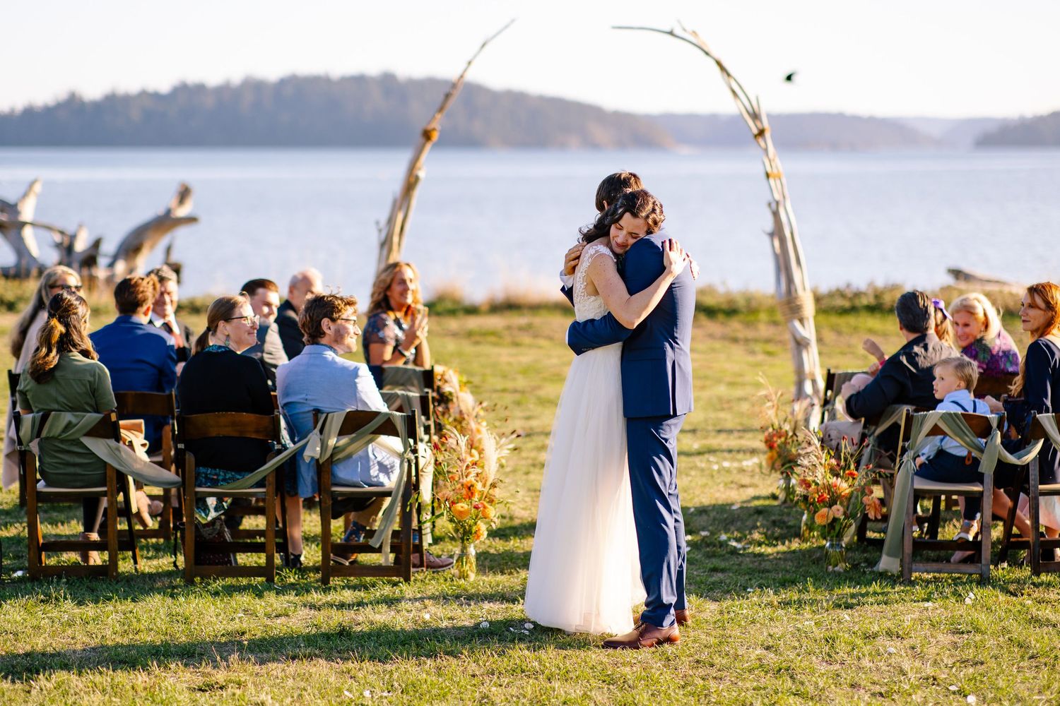 Bride and groom exchanging vows under a wooden arch by the ocean with guests seated nearby.