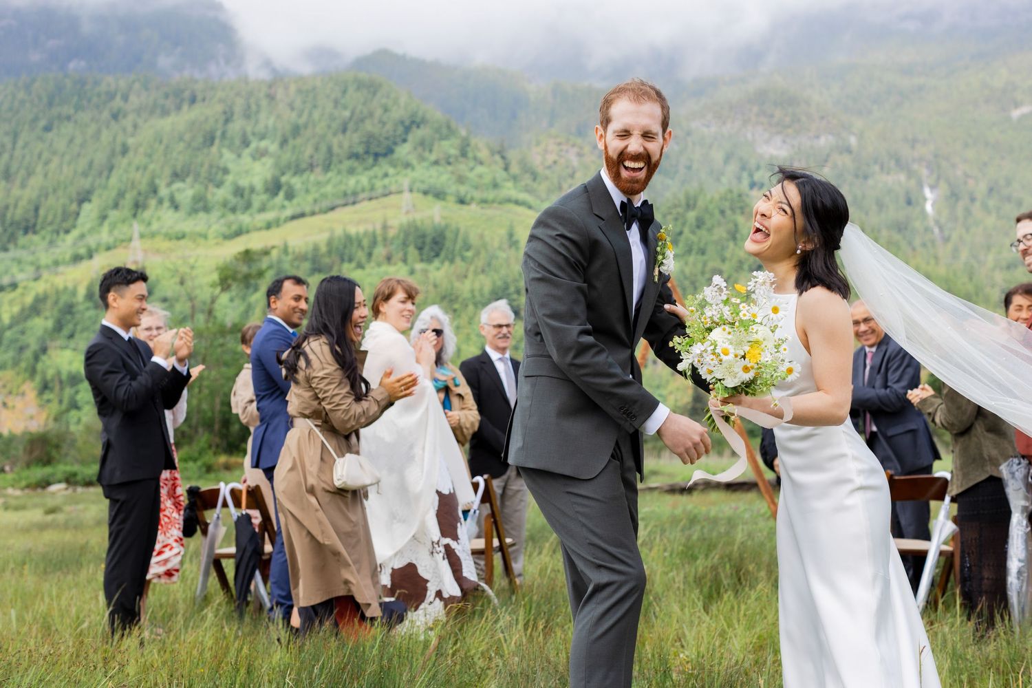 Wedding party celebrating in open field with mountain backdrop.