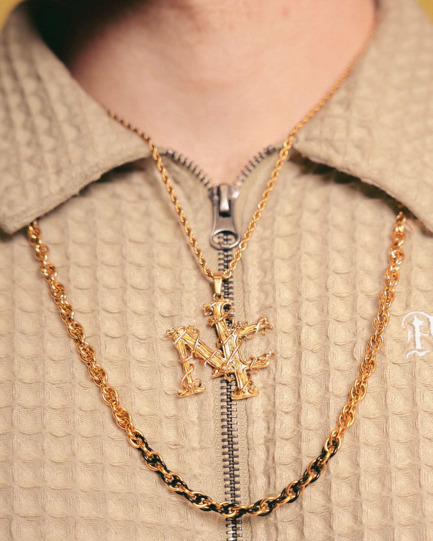 Gold cross pendant necklace layered with rope chain on model wearing beige textured zip-up jacket.