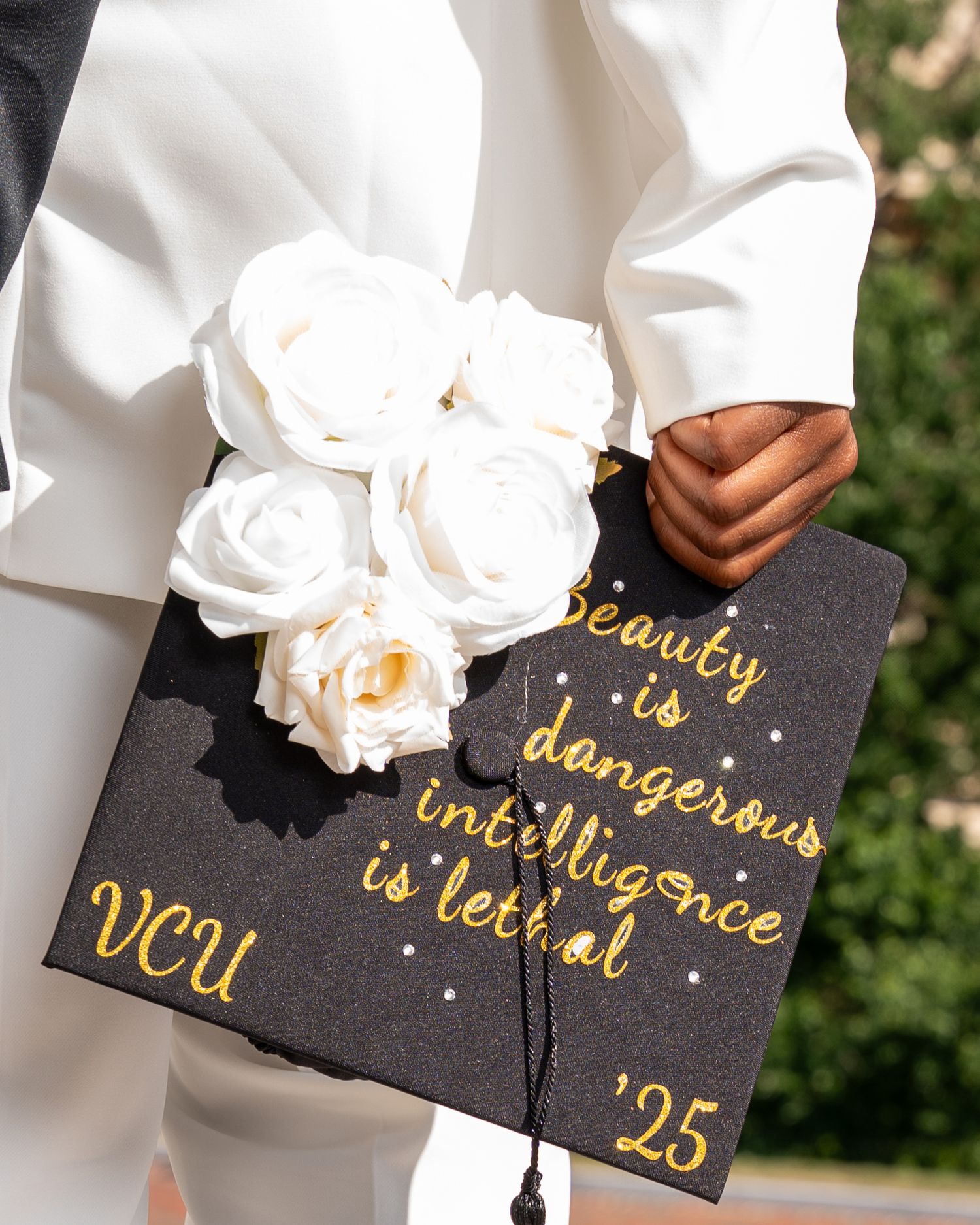 VCU 2025 graduate holds white roses behind a decorated black mortarboard cap with gold Beauty is dangerous intelligence is lethal quote.