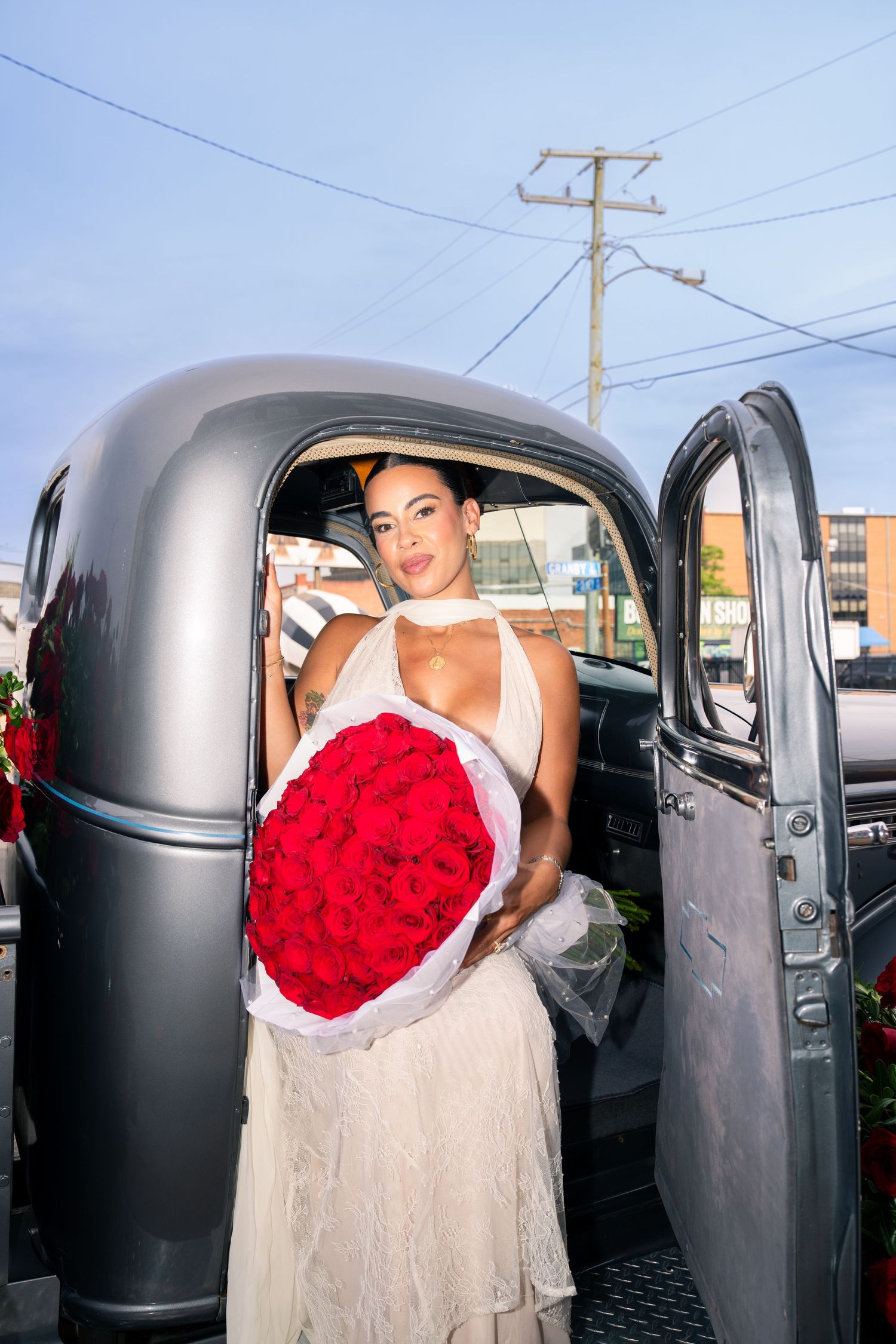 Woman in white dress holding large red rose bouquet beside a vintage black truck outdoors.