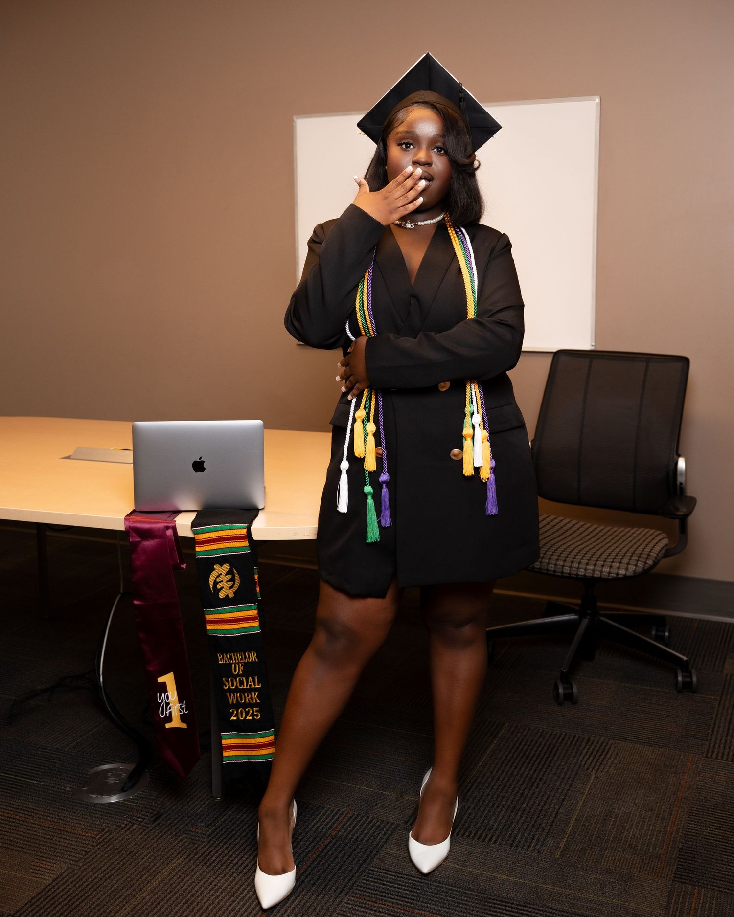 Graduate in black cap and gown with colorful honor cords poses in office, blowing a kiss in white heels.