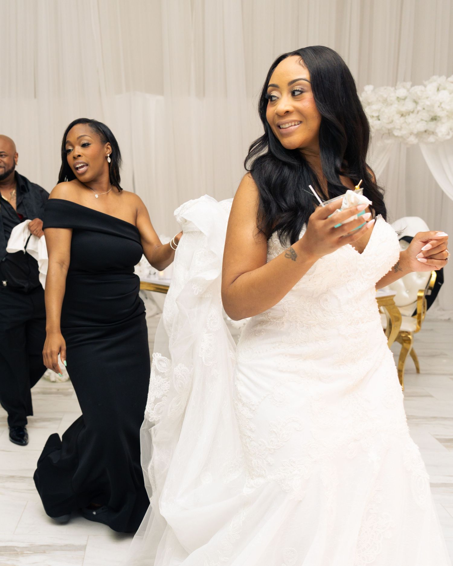 Bride in white wedding gown dancing joyfully with bridesmaid in black dress at elegant wedding reception.