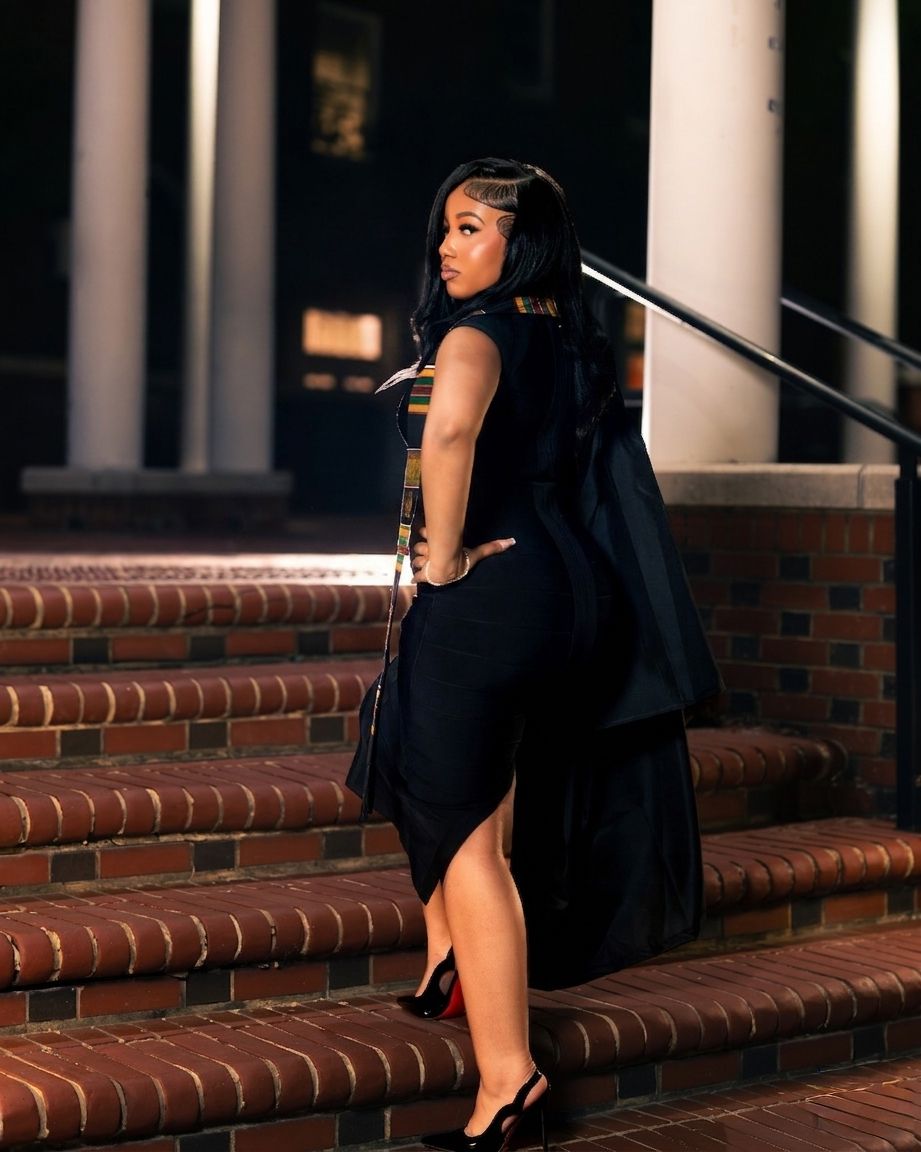 A woman in a black dress and heels poses on brick steps at night near white columns.
