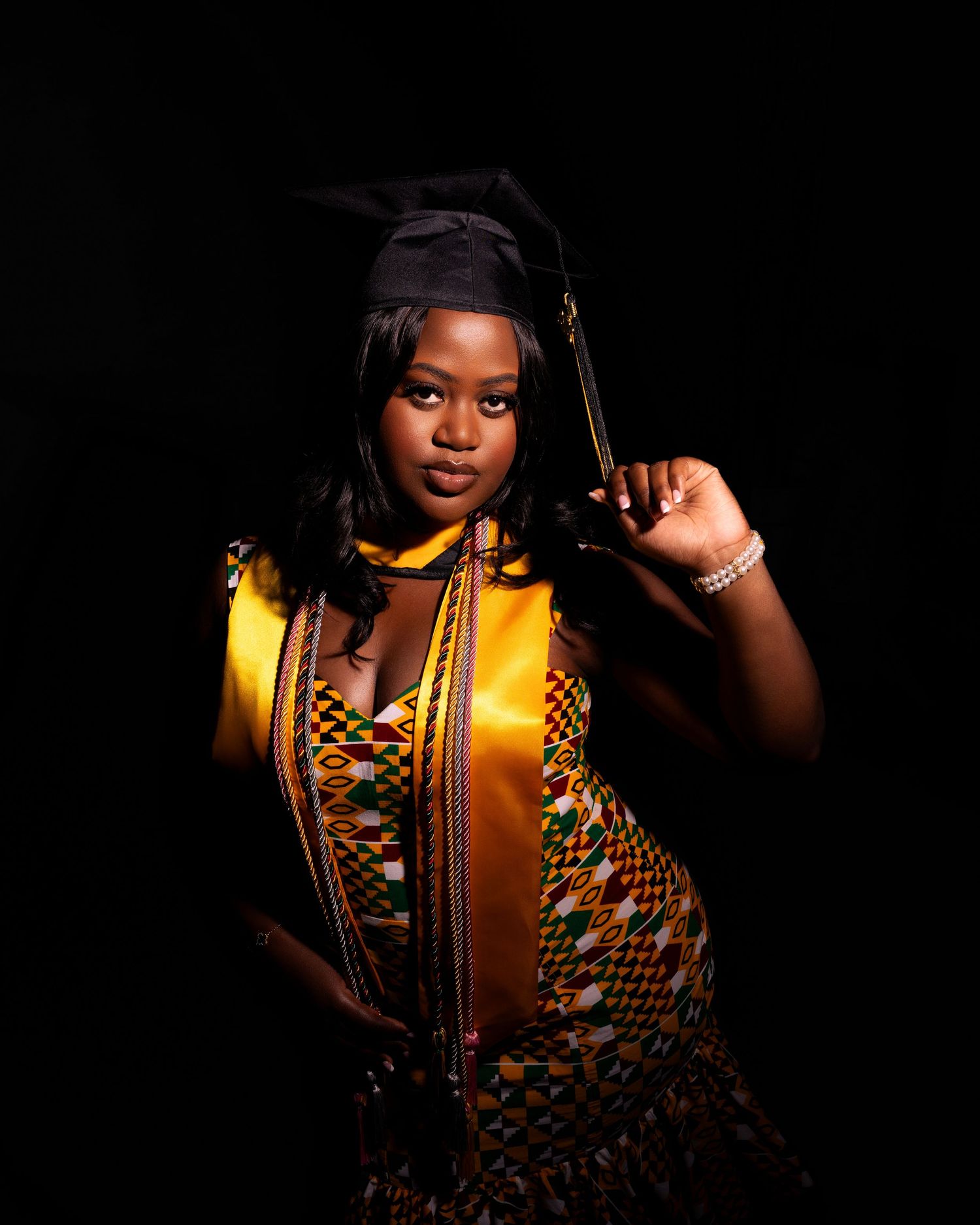 Young woman in graduation cap, gold sash, and kente cloth dress poses confidently against dark background.