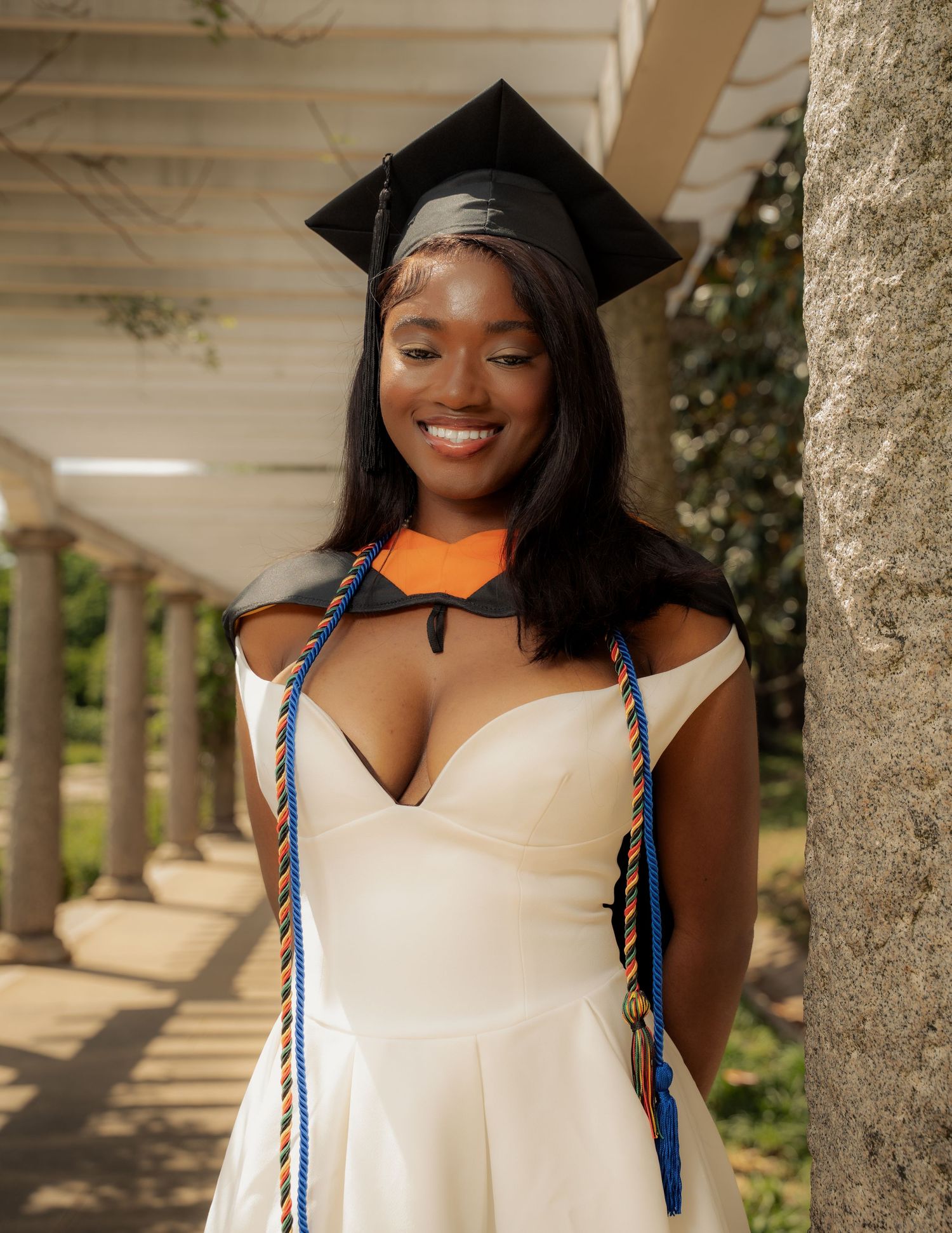 Smiling graduate in white dress and cap with colorful honor cords poses outdoors near stone columns.