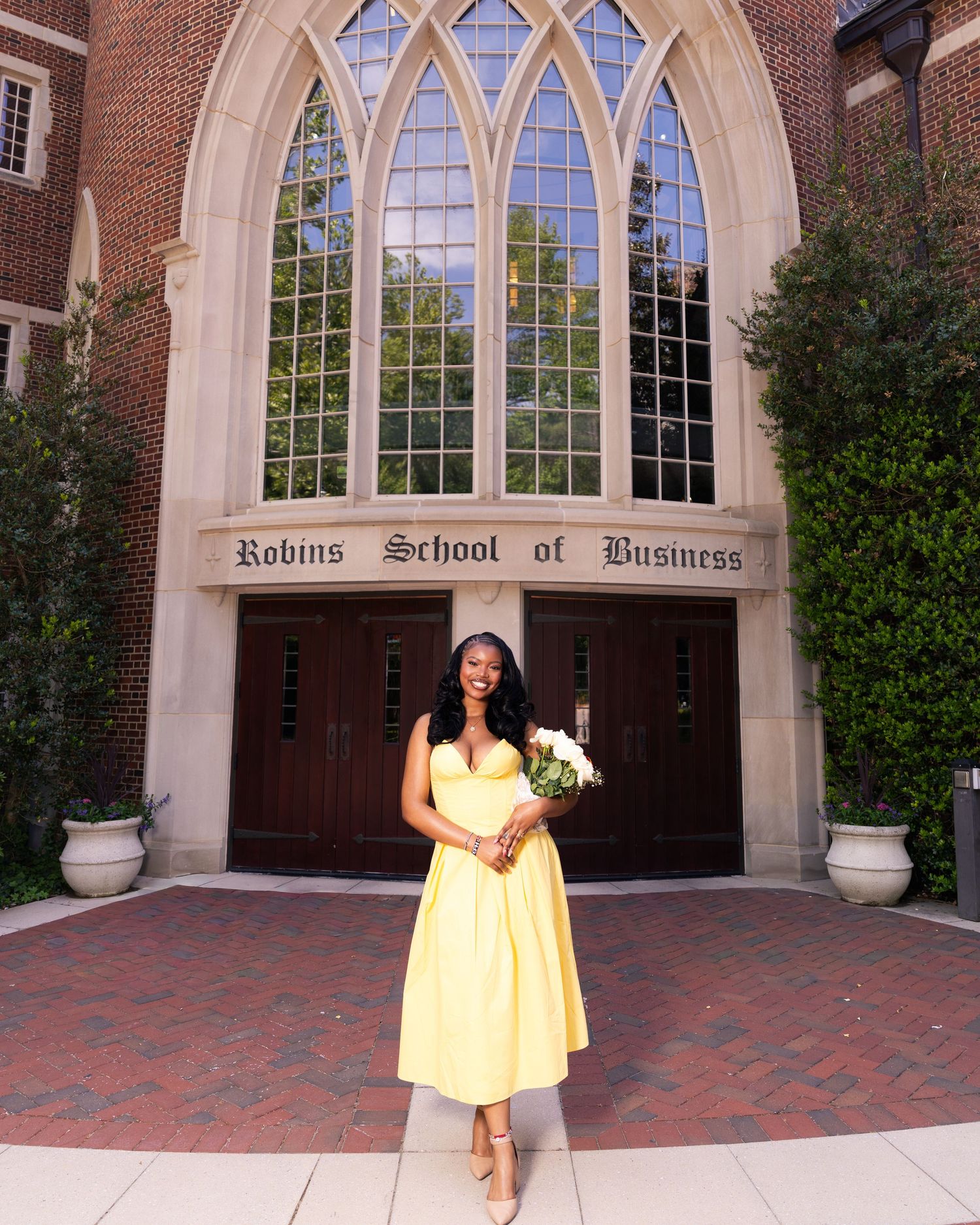 Woman in yellow dress holding flowers stands before Robins School of Business gothic brick building entrance.