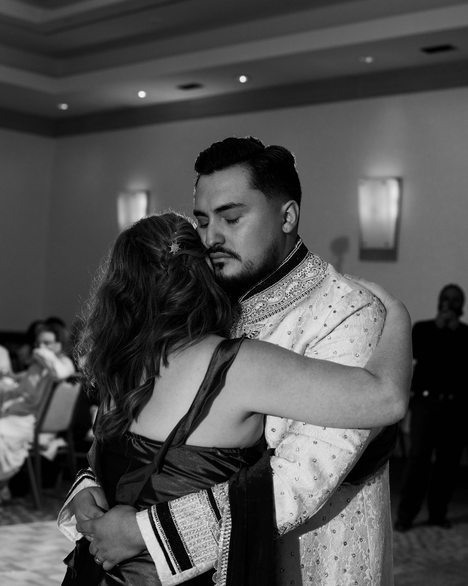 Black and white photo of a couple sharing an emotional embrace during a wedding dance in a banquet hall.