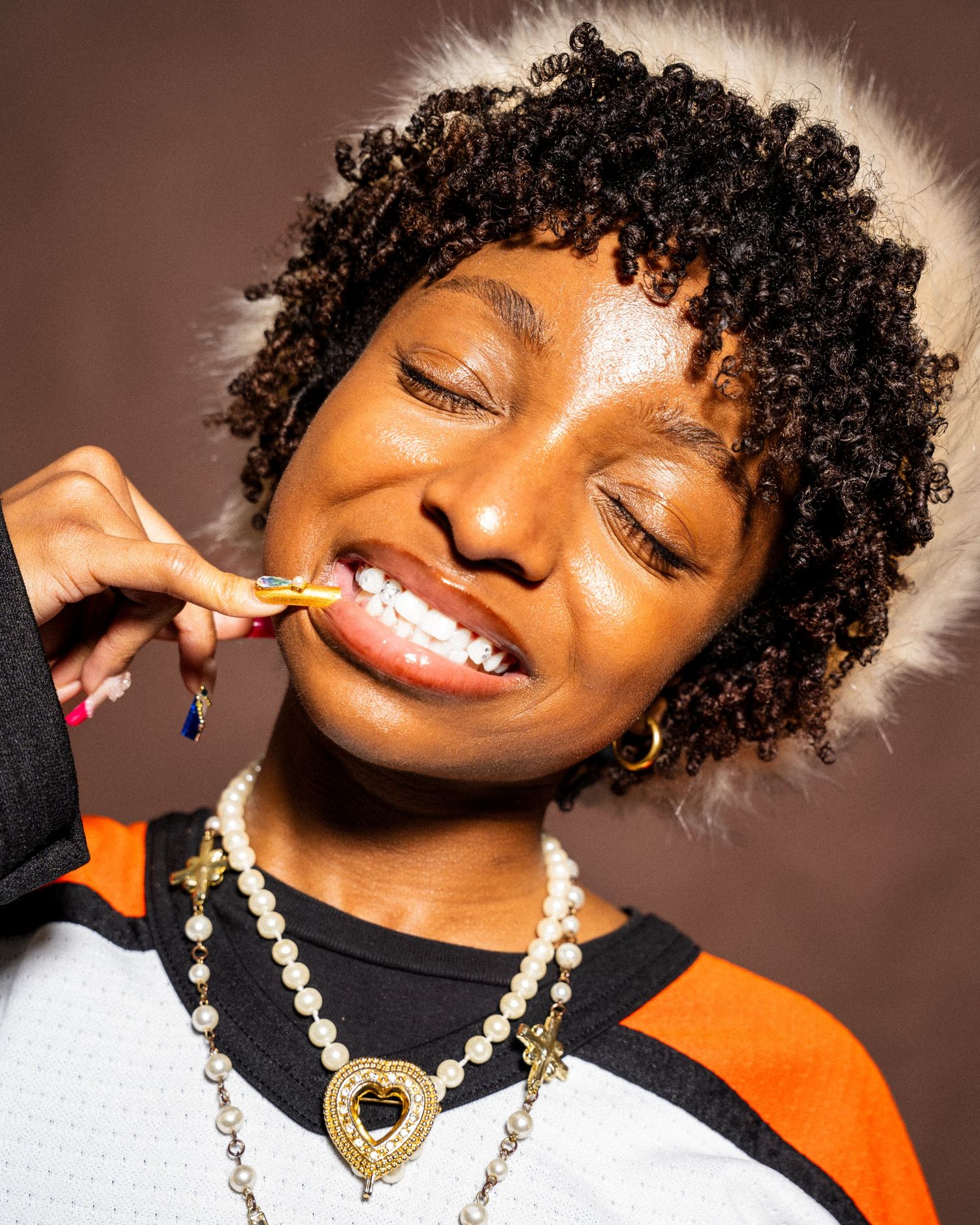 A young Black woman with curly hair brushes her teeth, wearing pearl and gold heart necklaces against a brown background.
