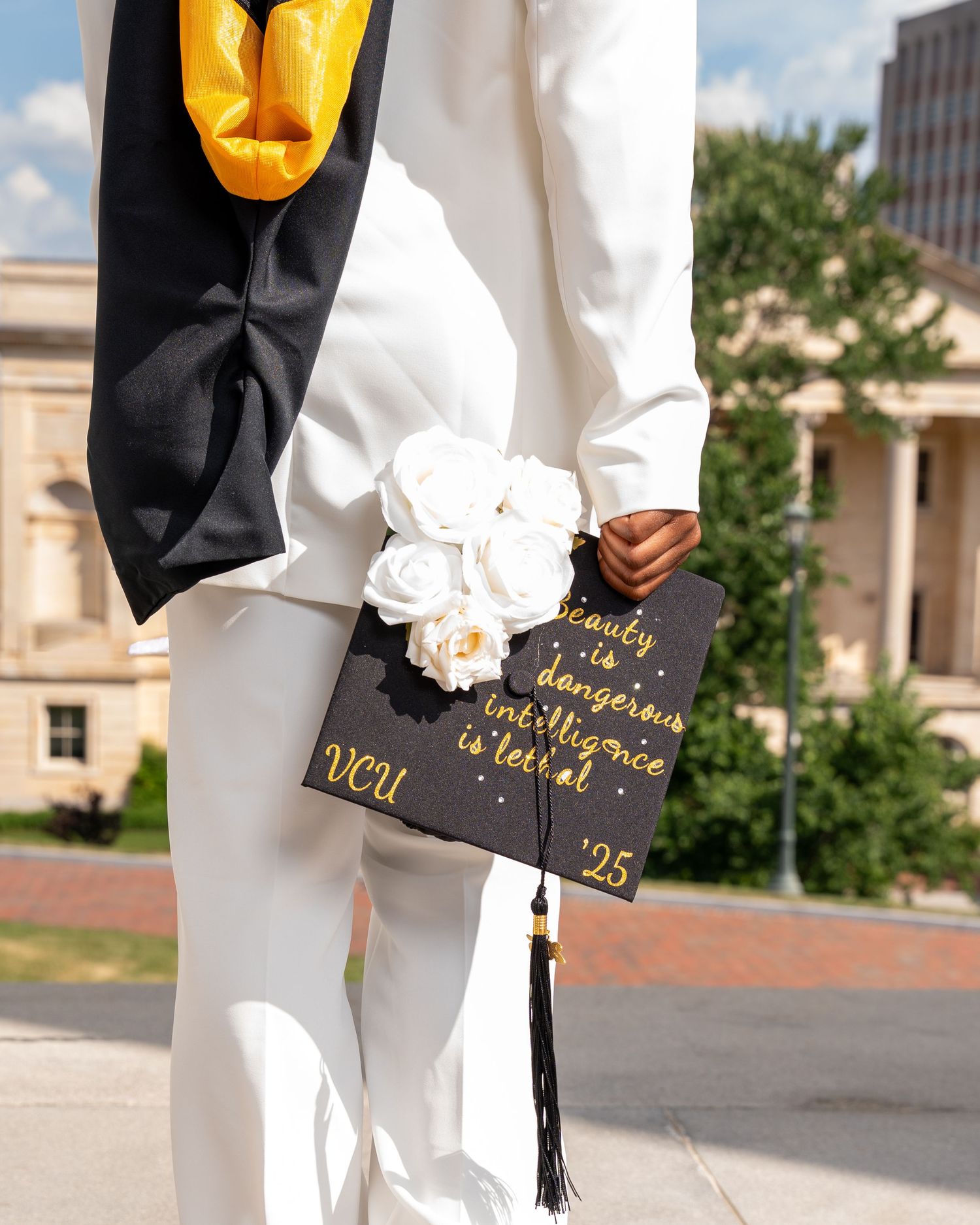 VCU 2025 graduate holding decorated black cap with white roses and gold text outdoors.