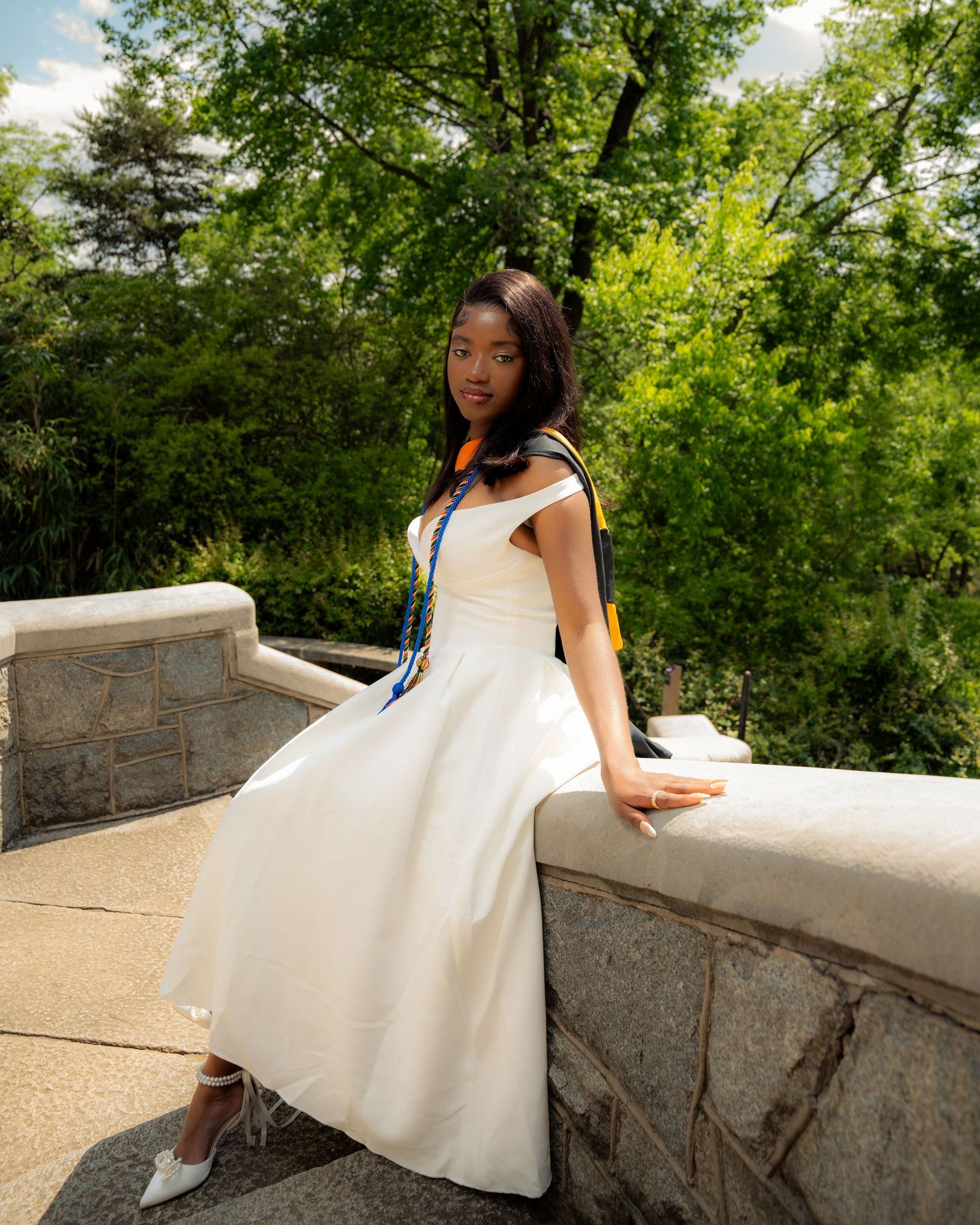Young woman in white midi dress with colorful sash poses on stone balustrade surrounded by lush green trees.