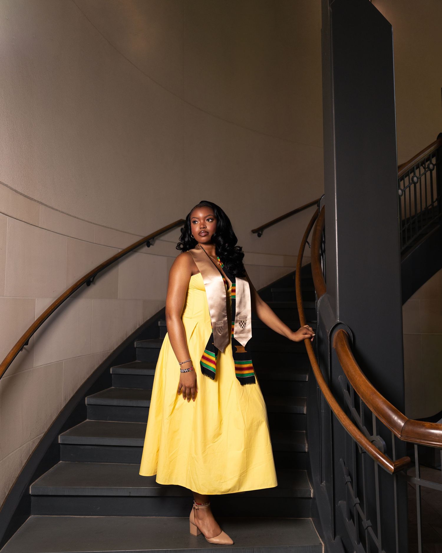 Graduate in yellow dress wearing kente cloth stole poses on elegant curved staircase during graduation celebration.