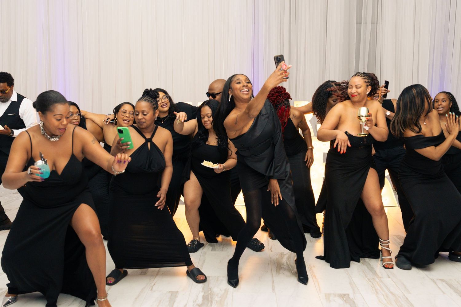 Group of women in black dresses dancing and taking selfies at a formal event or wedding reception.