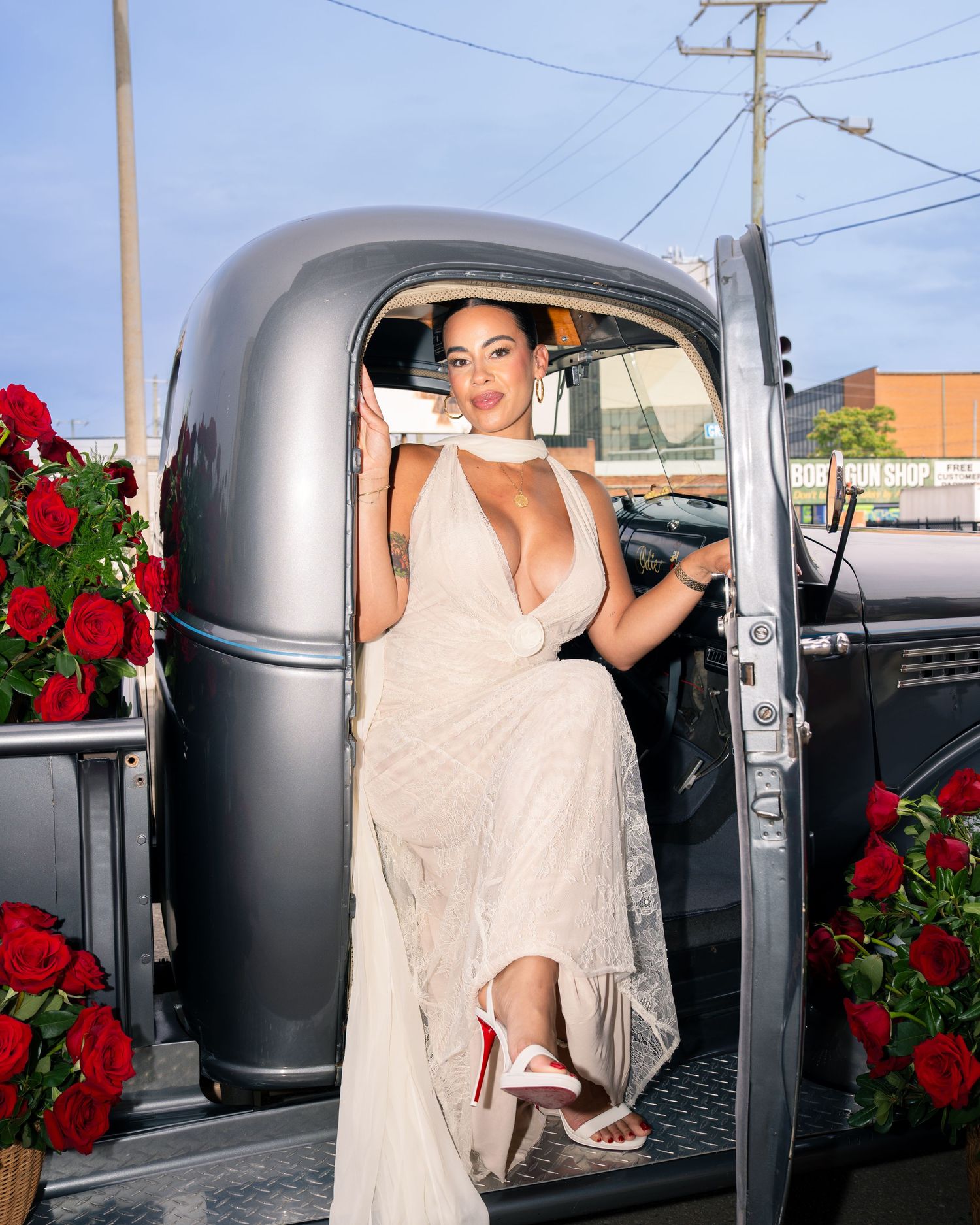Woman in elegant white halter gown poses in vintage truck doorway surrounded by red roses.