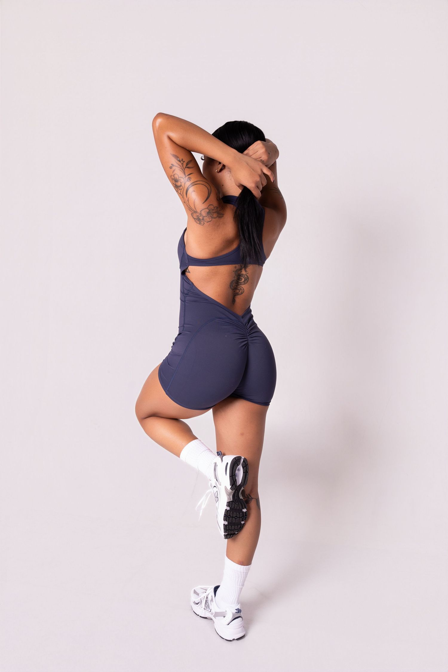 Woman in navy blue athletic crop top and shorts with white sneakers posing from behind, showing tattoos.