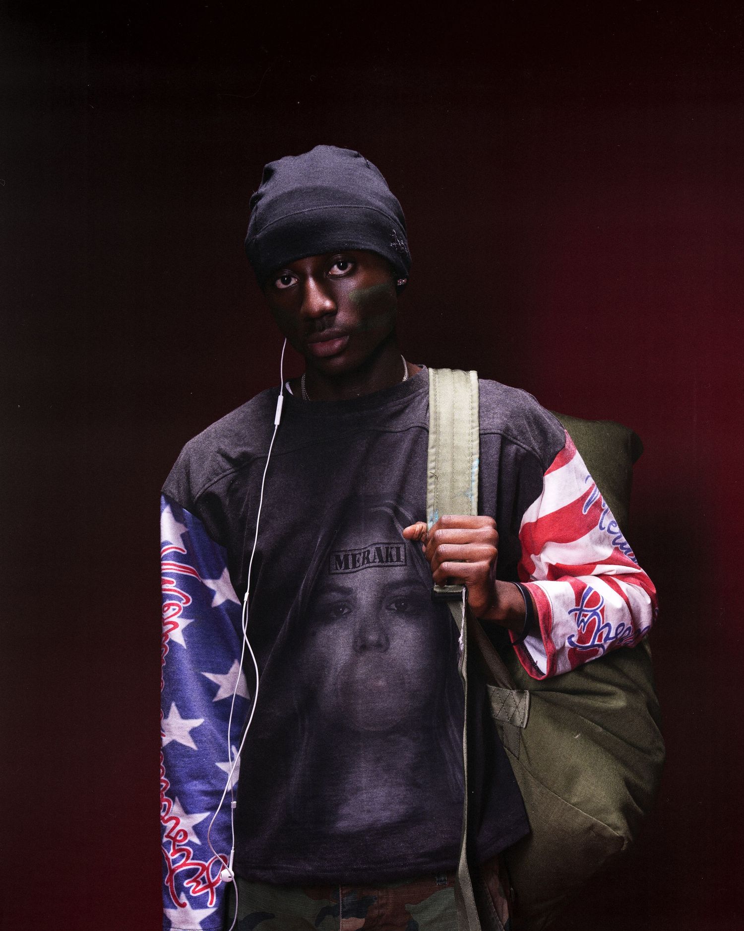Young man wearing American flag jacket, beanie hat, and graphic tee, carrying a green bag with earphones.