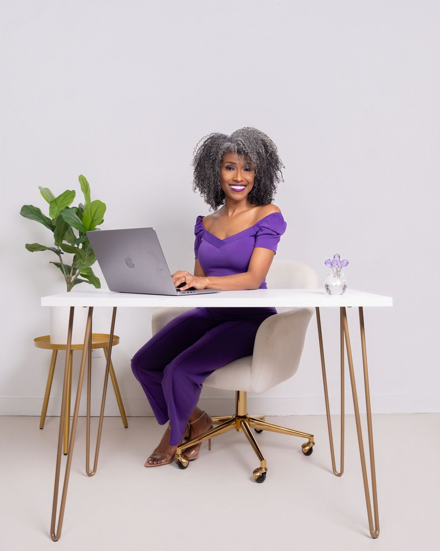 A smiling woman in a purple outfit sits at a white desk with a laptop in a bright, modern workspace.