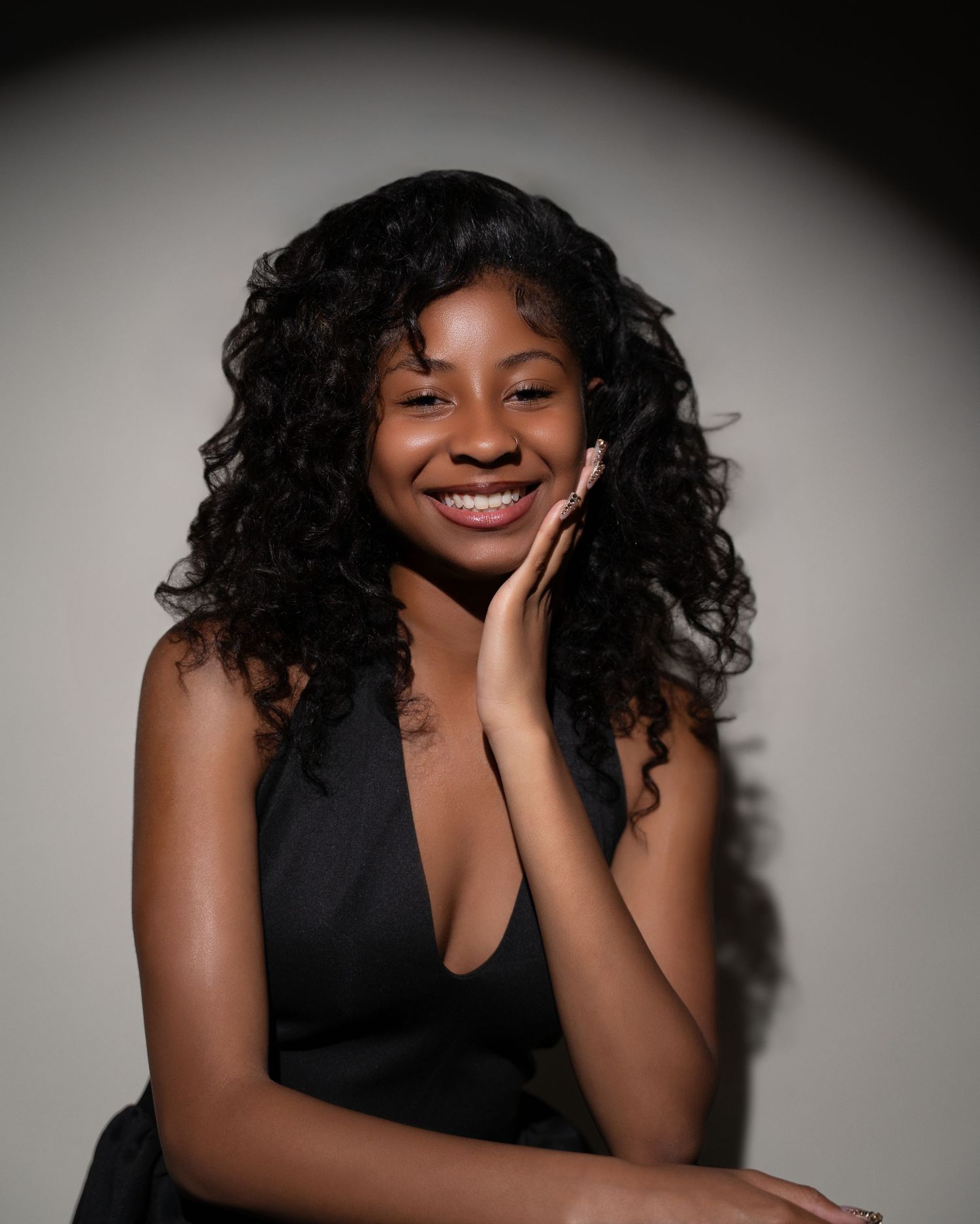 Young woman with curly black hair smiling warmly, wearing a black dress, posing gracefully against a dark background.