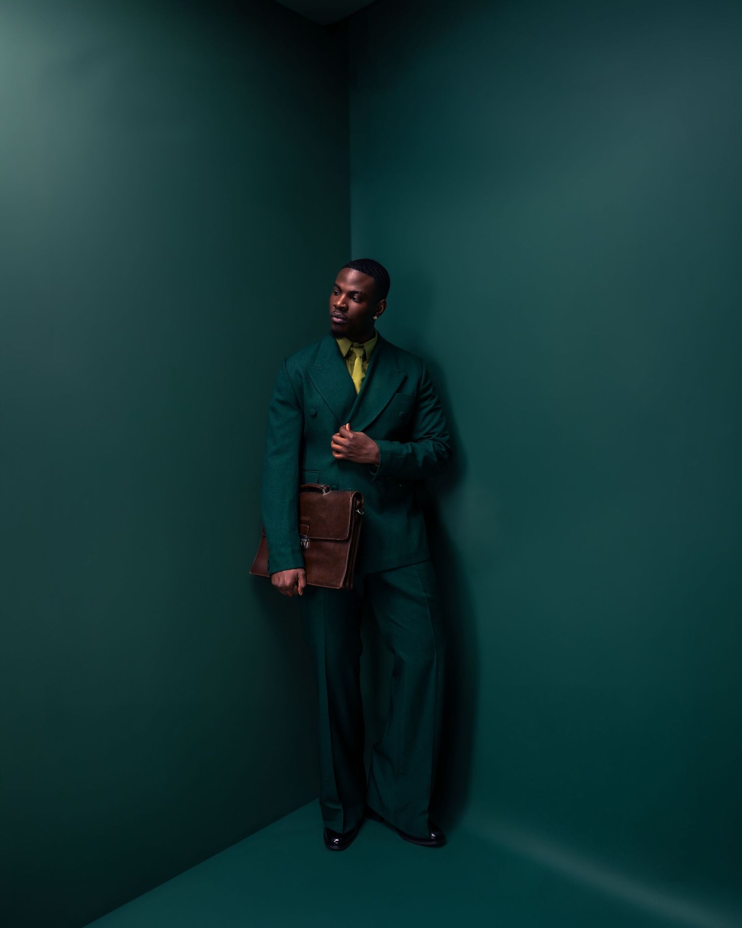 Man in dark green suit holding brown leather clutch, standing in teal corner for a stylish editorial look.