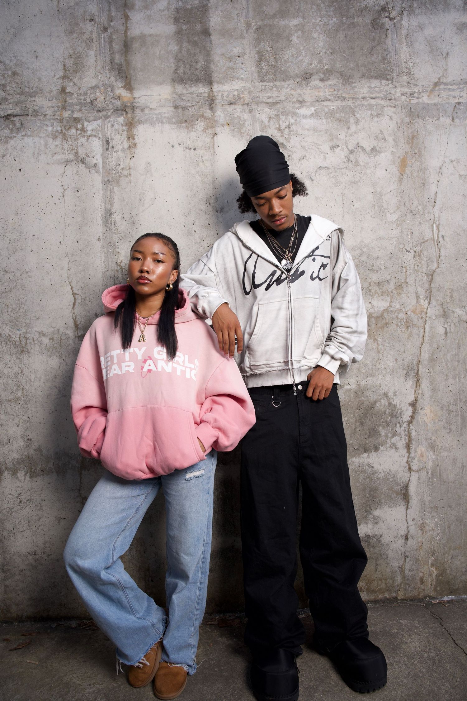 Two young people in streetwear pose against a concrete wall, wearing oversized hoodies and baggy jeans.